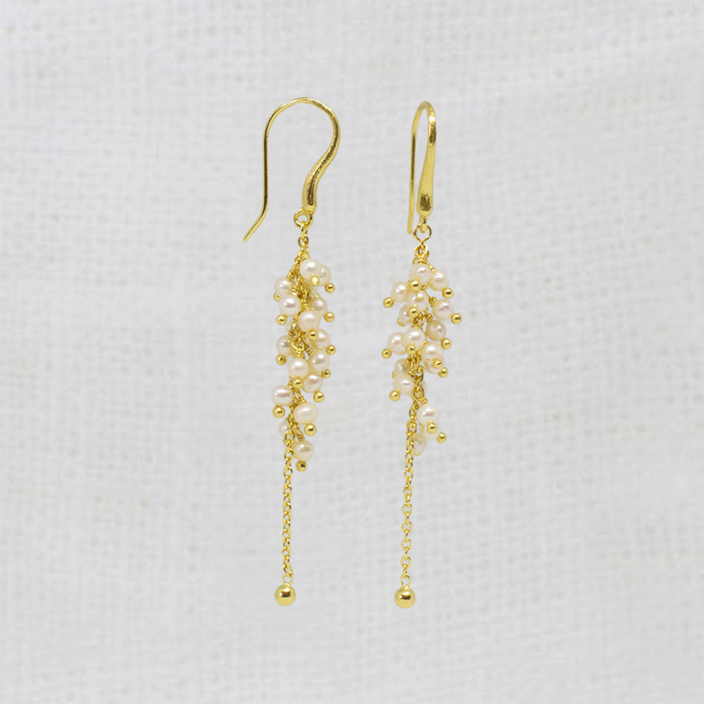 A pair of gold dangle earrings with clusters of small white pearls and gold beads, hanging against a textured white fabric background.