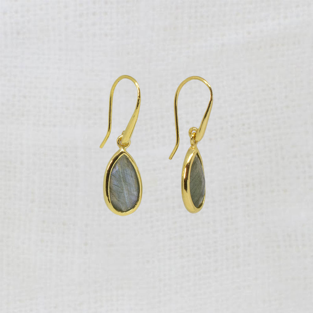A pair of gold drop earrings with teardrop-shaped gray-green labradorite stones, displayed on a textured white fabric background.