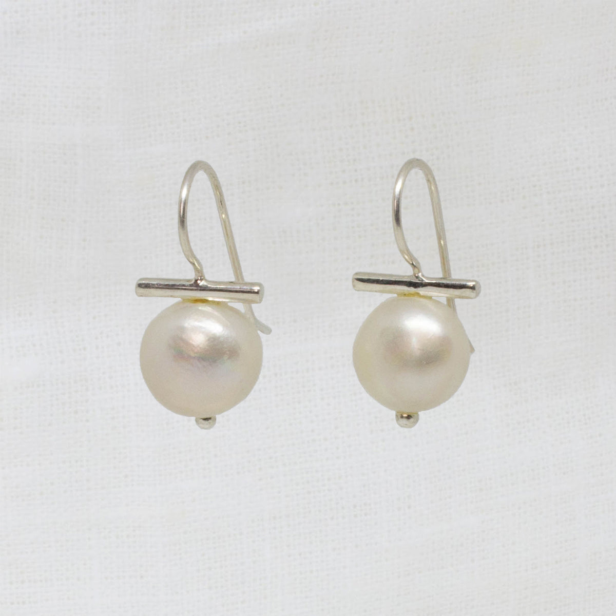 A pair of silver fishhook earrings with a small horizontal bar and a single round white pearl dangling below, shown on a white textured fabric background.