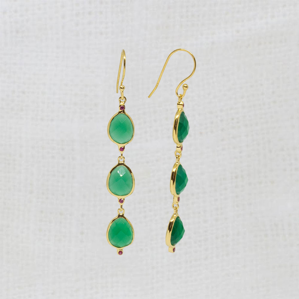 A pair of gold drop earrings with three green teardrop gemstones and tiny red accent stones, displayed on a textured white fabric background.