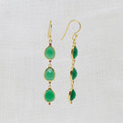 A pair of gold drop earrings with three green teardrop gemstones and tiny red accent stones, displayed on a textured white fabric background.