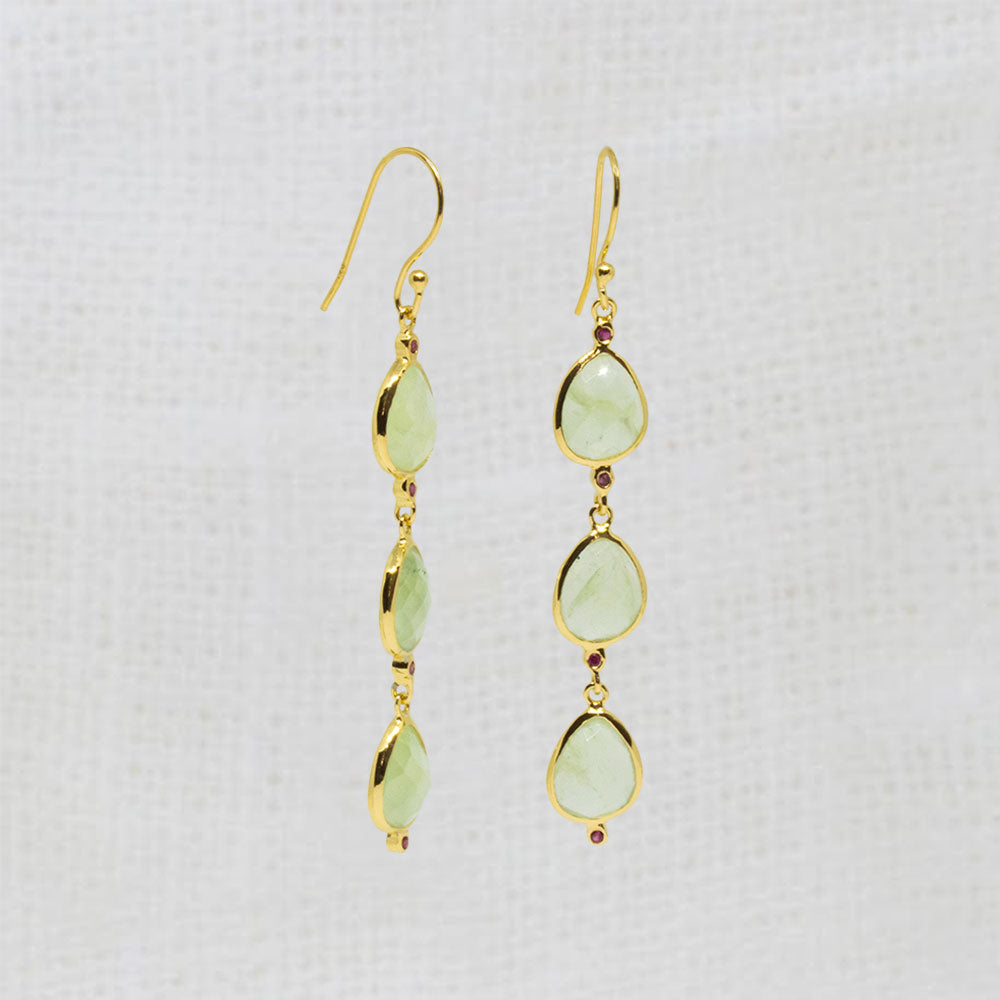 A pair of gold dangle earrings with three light green teardrop-shaped gemstones and tiny red accent beads, shown on a textured white fabric background.