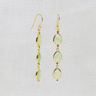 A pair of gold dangle earrings with three light green teardrop-shaped gemstones and tiny red accent beads, shown on a textured white fabric background.