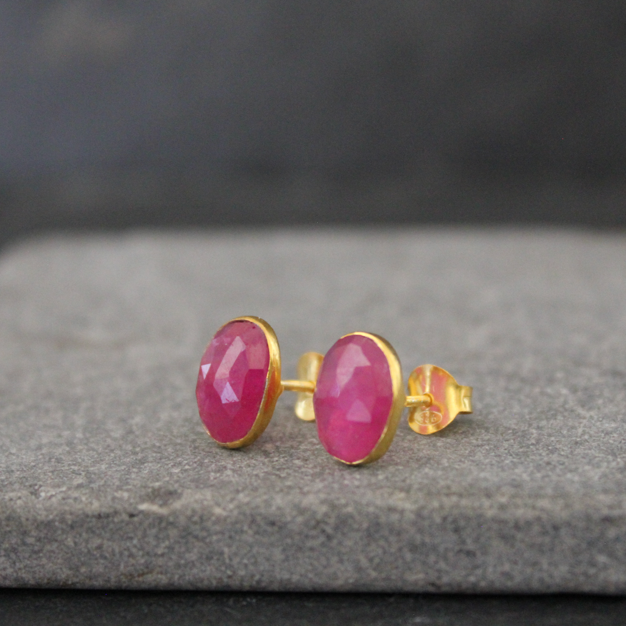 A pair of gold stud earrings with oval faceted pink ruby gemstones, resting on a textured gray stone surface.