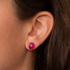 A close-up of a person's ear wearing a small oval stud earring with a vivid pink cabochon stone and a gold-tone setting.