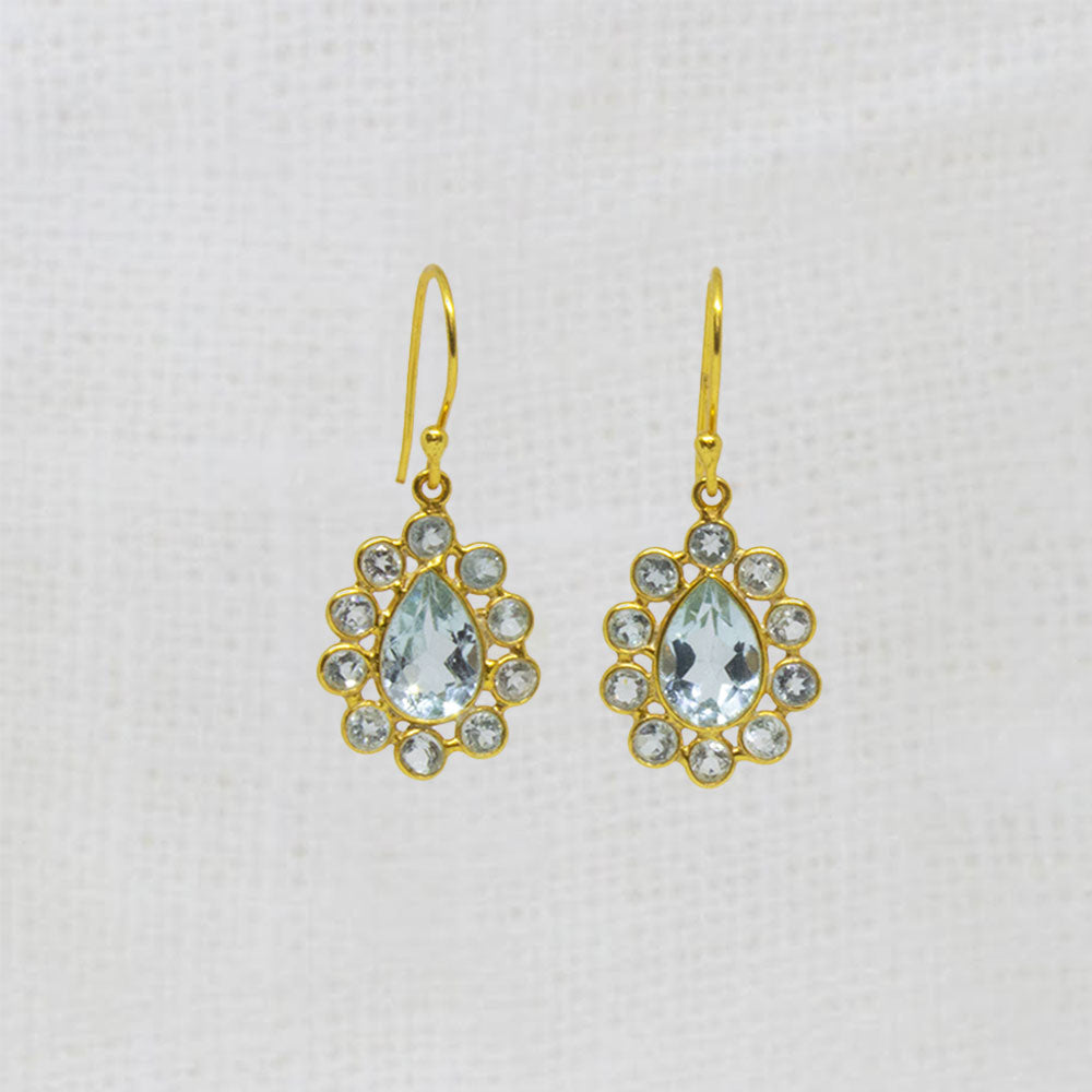 A pair of gold drop earrings featuring a central pear-shaped light blue topaz gemstone surrounded by small blue topaz round stones, displayed on a textured off-white fabric background.