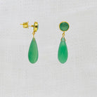 A pair of gold drop earrings featuring faceted green teardrop-shaped stones, shown against a textured white fabric background.