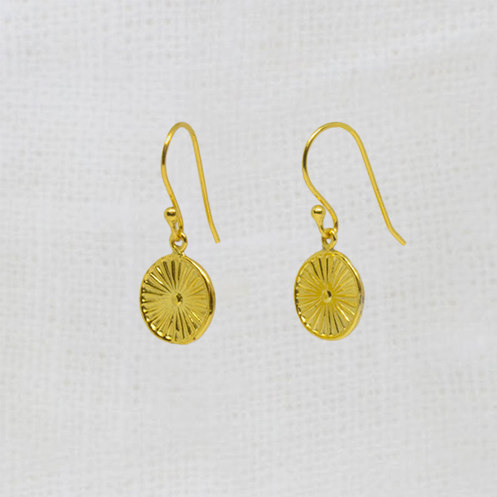 A pair of gold dangle earrings with textured circular disc pendants, displayed on a white textured fabric background.