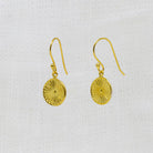 A pair of gold dangle earrings with textured circular disc pendants, displayed on a white textured fabric background.