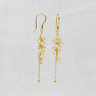 A pair of gold dangle earrings with clusters of small white pearls and gold beads, hanging against a textured white fabric background.
