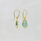A pair of gold drop earrings with teardrop-shaped light green gemstone pendants, displayed against a textured white fabric background.