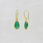 A pair of gold teardrop dangle earrings with green cabochon gemstones, displayed on a textured white fabric background.