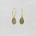 A pair of gold teardrop dangle earrings with gray-green labradorite stones, displayed on a textured white fabric background.