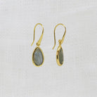 A pair of gold drop earrings with teardrop-shaped gray-green labradorite stones, displayed on a textured white fabric background.