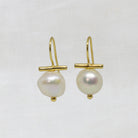 A pair of gold drop earrings featuring a small horizontal gold bar and a single irregular white baroque pearl hanging below, displayed on a textured white fabric background.