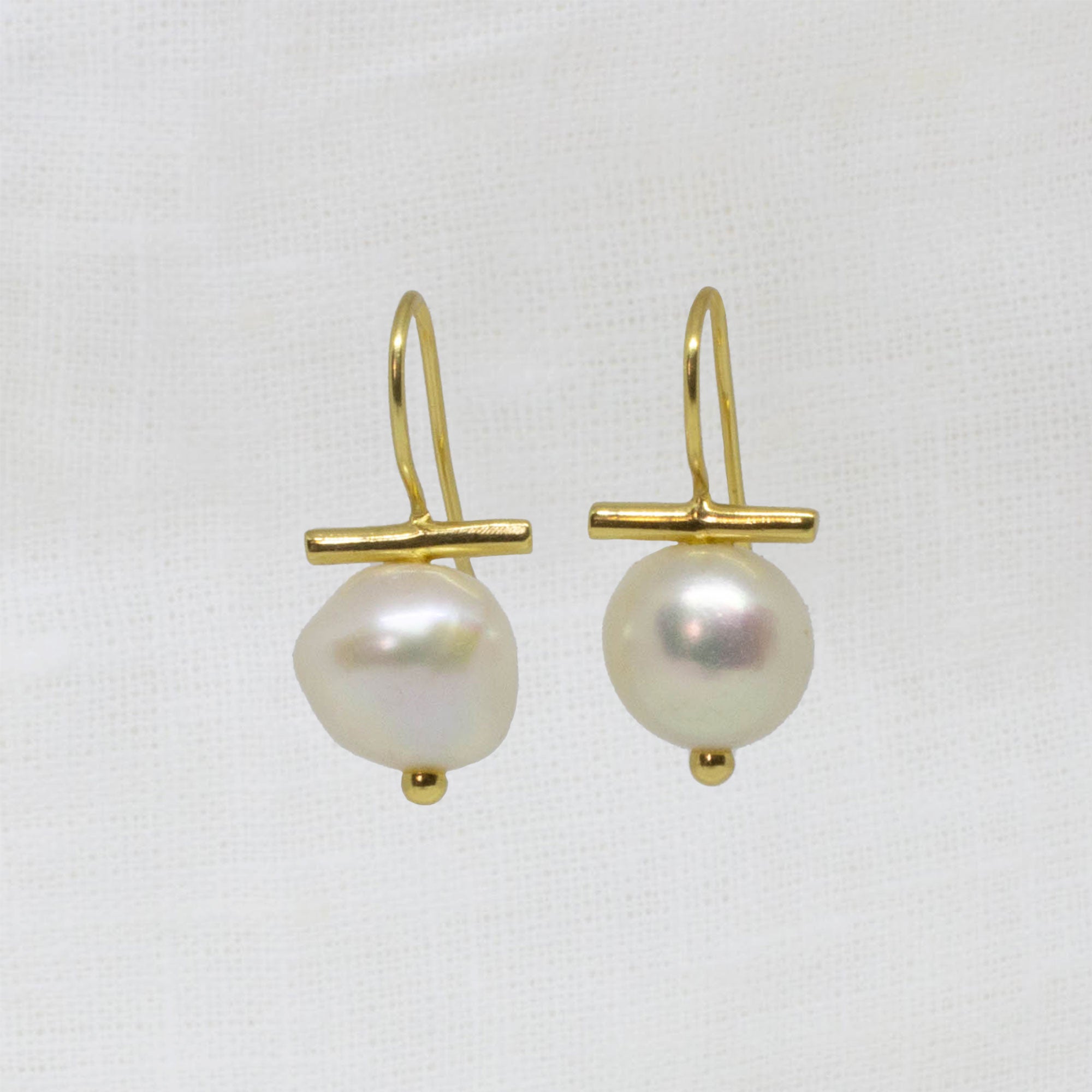 A pair of gold drop earrings featuring a small horizontal gold bar and a single irregular white baroque pearl hanging below, displayed on a textured white fabric background.