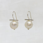 A pair of silver fishhook earrings with a small horizontal bar and a single round white pearl dangling below, shown on a white textured fabric background.