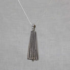 A silver pendant necklace featuring a geometric, cone-shaped design with vertical rows of small clear stones, suspended from a delicate silver chain against a textured light gray background.